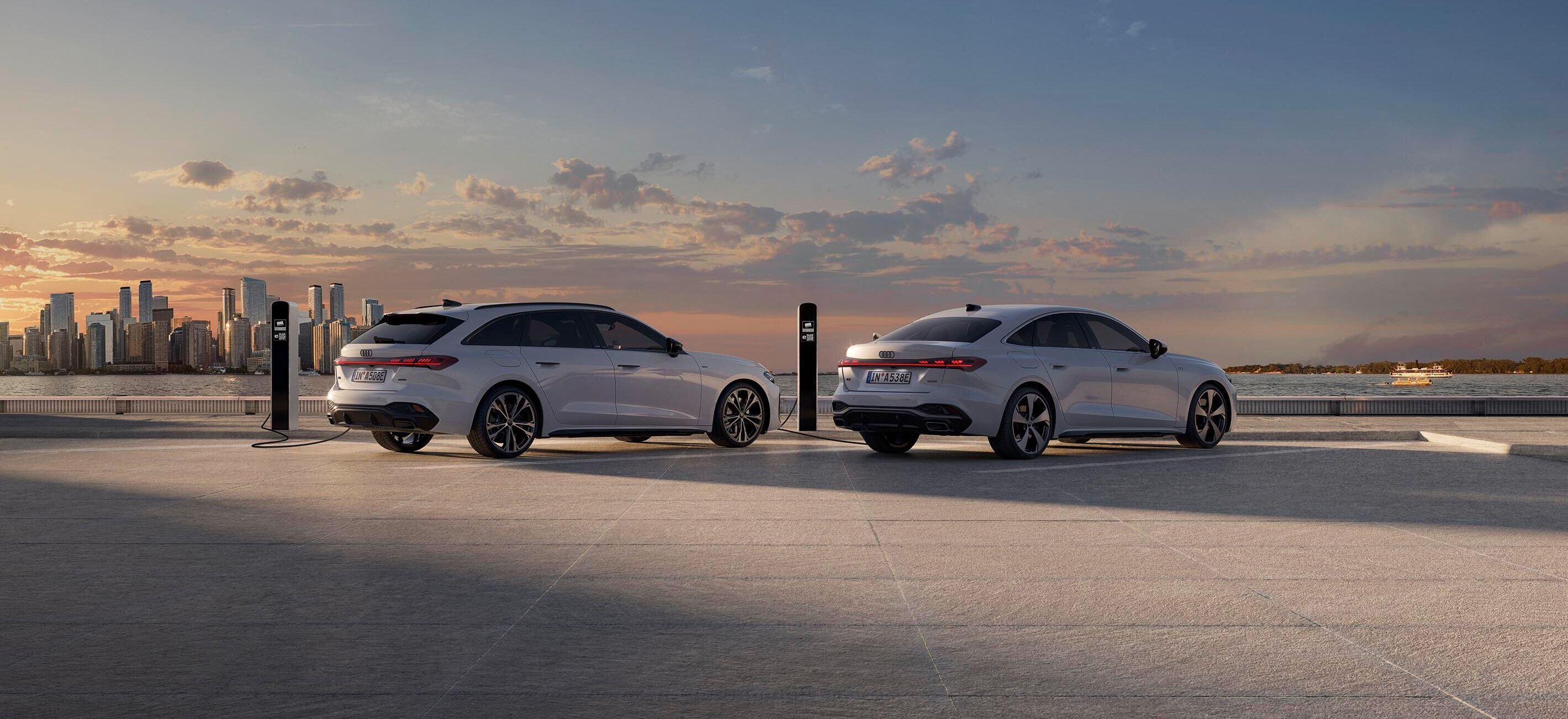 Zwei weiße Audi A5 e-tron Fahrzeuge, ein Kombi und eine Limousine, stehen nebeneinander auf einer offenen Fläche mit Blick auf eine Stadt bei Sonnenuntergang