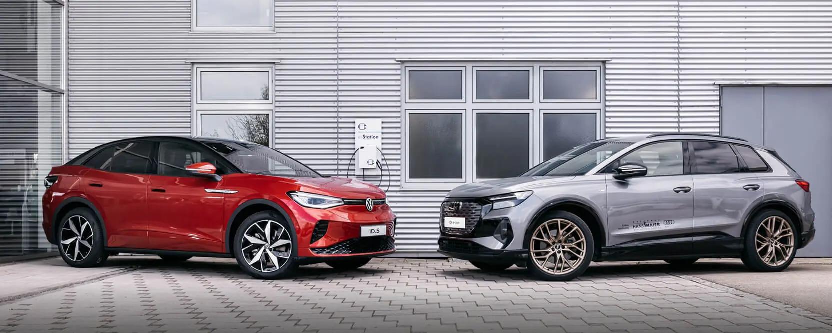 ein roter VW ID.5 und ein grauer Audi Q4 e-tron stehen vor einer grauen gewellten Metallwand, im Hintergrund ist eine Ladestation zu sehen