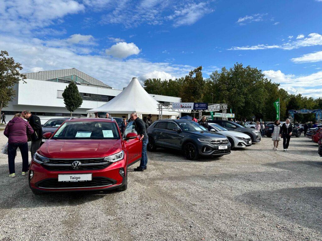 Außenaufnahme einer Automobilausstellung mit mehreren geparkten Autos, vorne ein roter Volkswagen Taigo, im Hintergrund ein weißes Zelt und Besucher