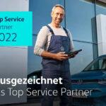Serviceberater mit blauer Latzhose von VW steht vor einem VW Gebäude, Text mit der Aufschrift 'Ausgezeichnet als Top Service Partner 2022'