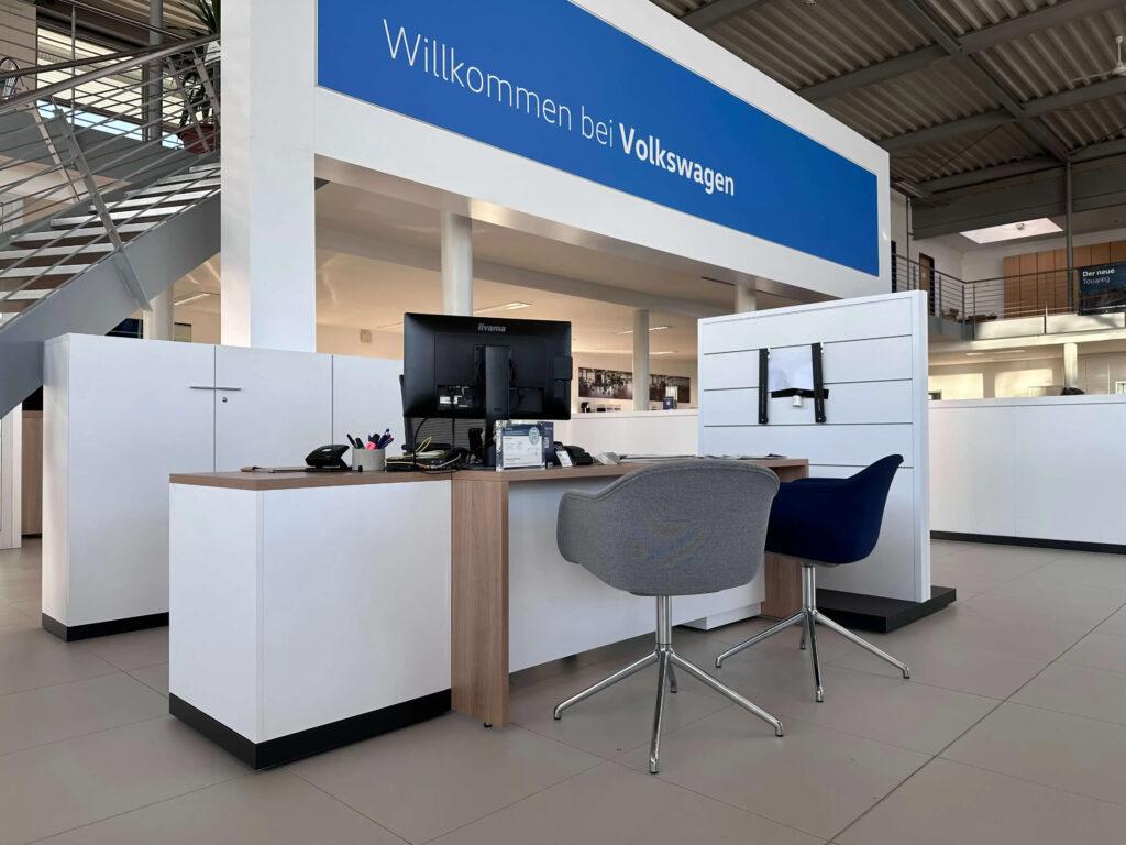 Empfangstresen mit zwei Stühlen und zwei Monitoren in einem hellen Volkswagen-Autohaus, darüber ein blaues Schild mit der Aufschrift 'Willkommen bei Volkswagen'.