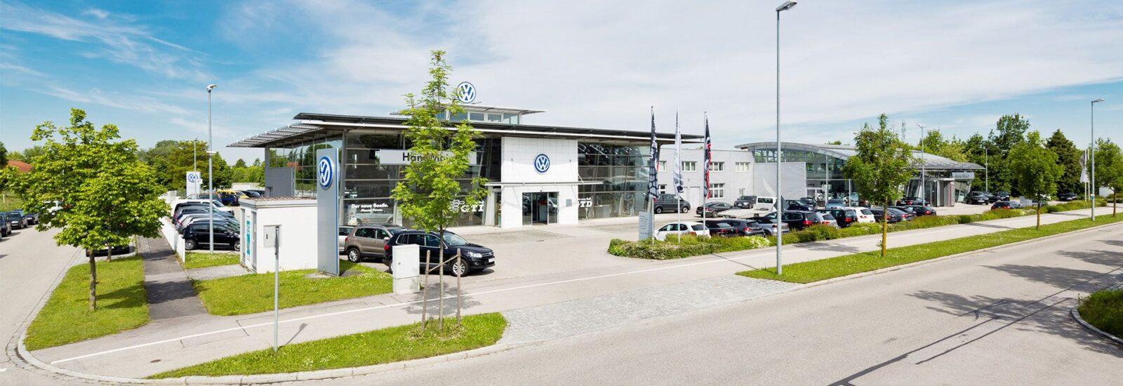Außenaufnahme eines Volkswagen-Autohauses mit Glasfassade, mehreren geparkten Autos und Bäumen entlang der Straße bei blauem Himmel.