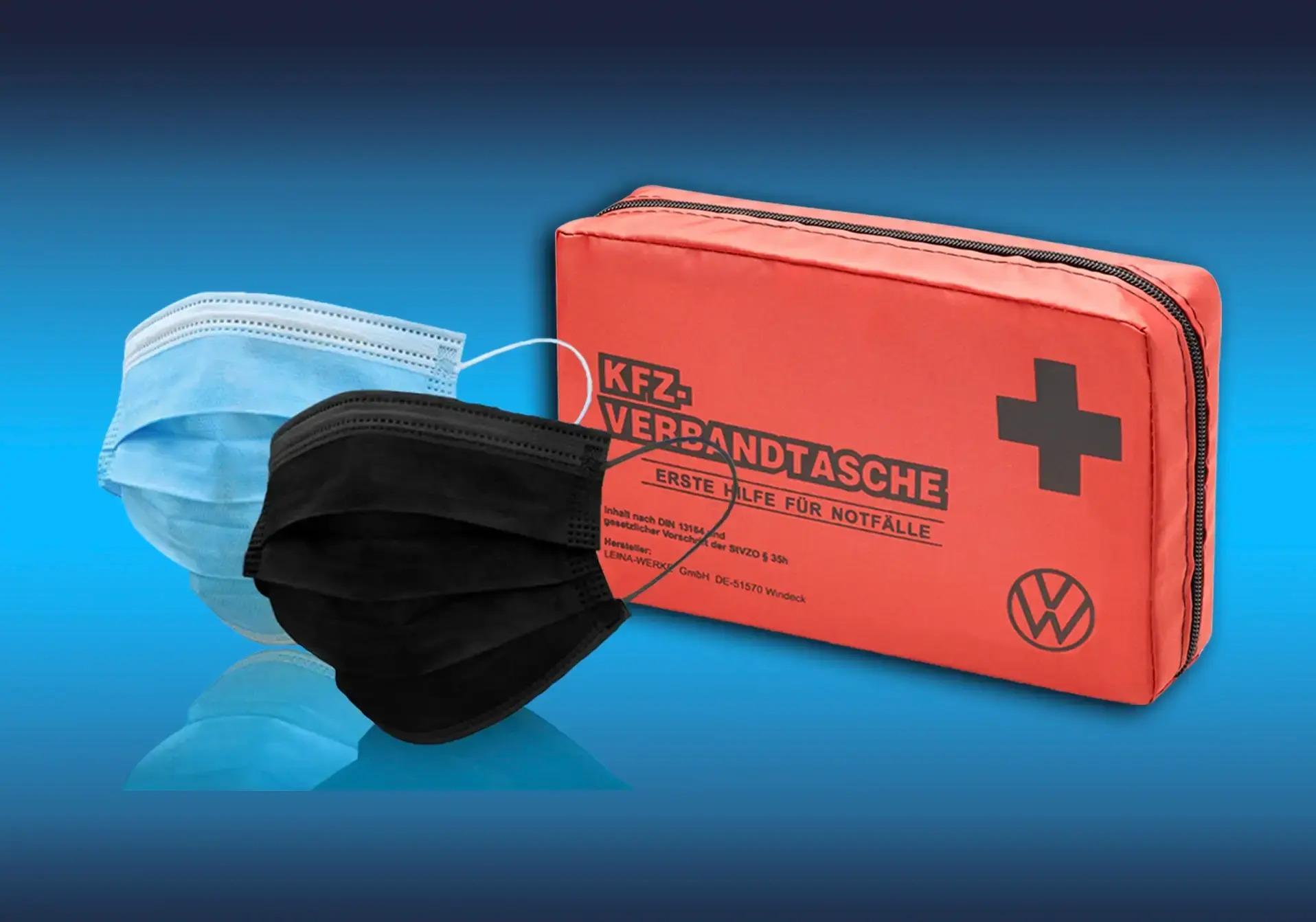 eine blaue und eine schwarze Atemschutzmaske vor einer roten Kfz-Verbandtasche auf blauem Hintergrund