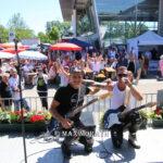 Zwei Musiker mit E-Gitarren knien auf einer Bühne vor einem Publikum im Freien, umgeben von Blumen und Sonnenschirmen.