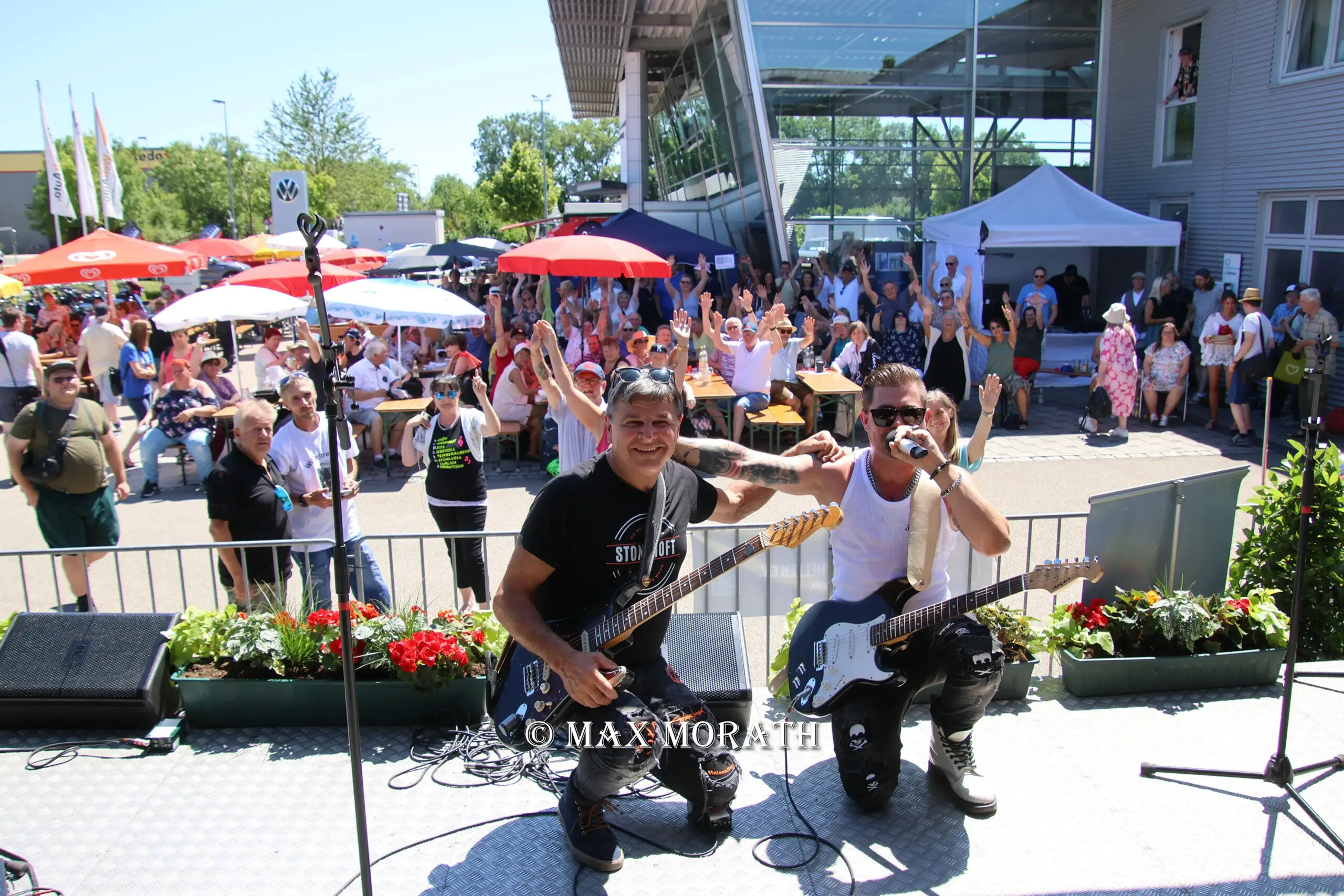Zwei Musiker mit E-Gitarren knien auf einer Bühne vor einem Publikum im Freien, umgeben von Blumen und Sonnenschirmen.