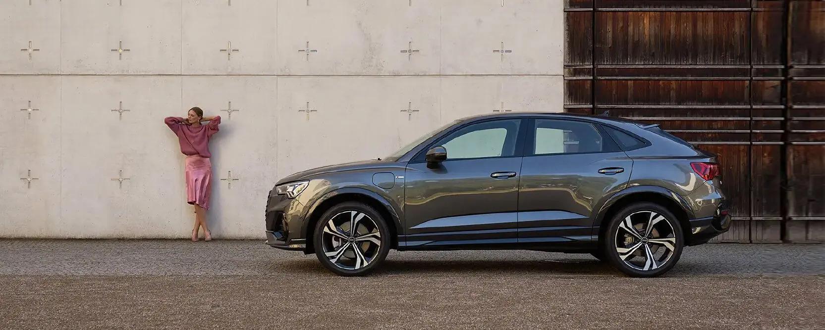 Audi Leasingangebote für Privatkunden 1 Frau mit pinkem Satinrock und dunkelpinken Pullover lehnt an einer Wand vor einem grauen Audi Q4