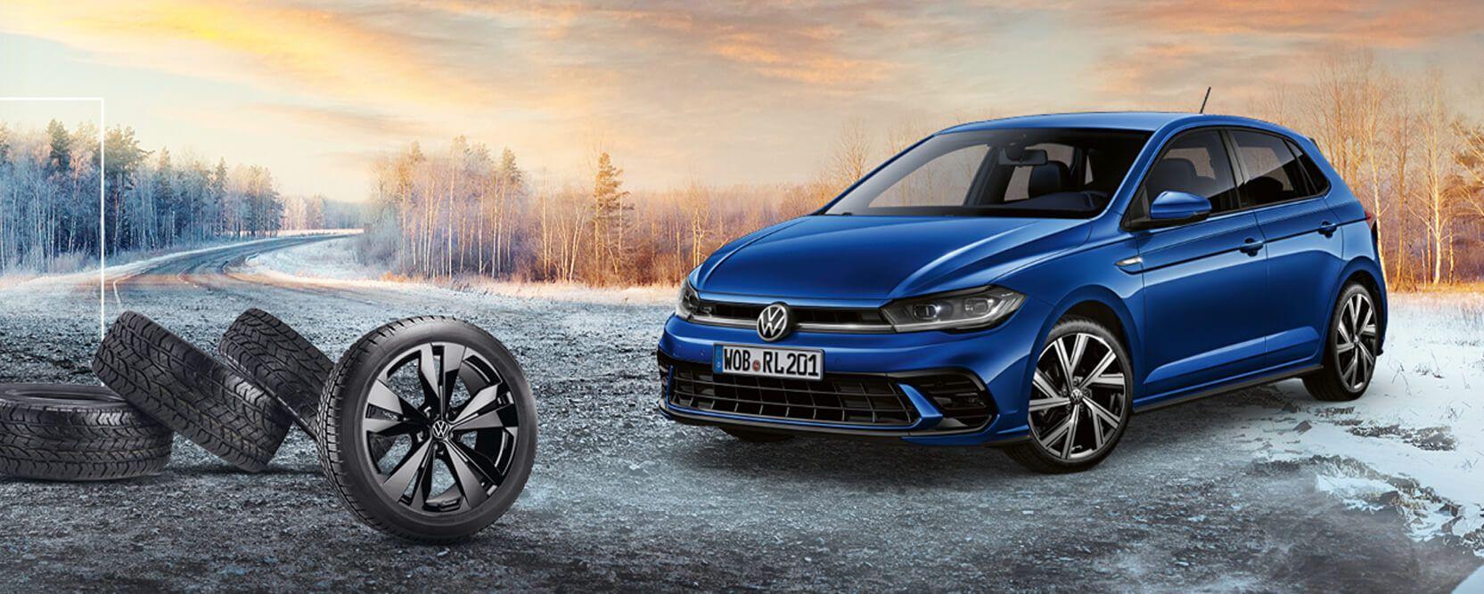 Blauer Volkswagen Polo auf winterlicher Straße, daneben vier Autoreifen, zwei davon mit Felgen, im Hintergrund herbstliche Bäume und Sonnenuntergang.