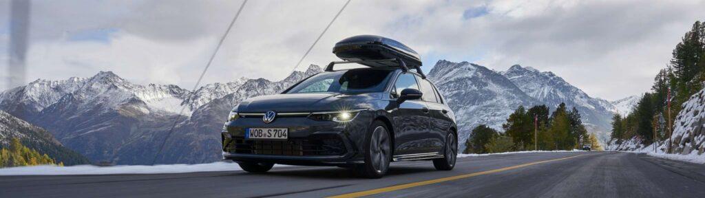 Reifenwechsel-Aktionstage im Herbst 30 Schwarzer VW Golf mit Dachbox fährt auf Bergstraße, schneebedeckte Berge und Nadelbäume im Hintergrund, bewölkter Himmel.