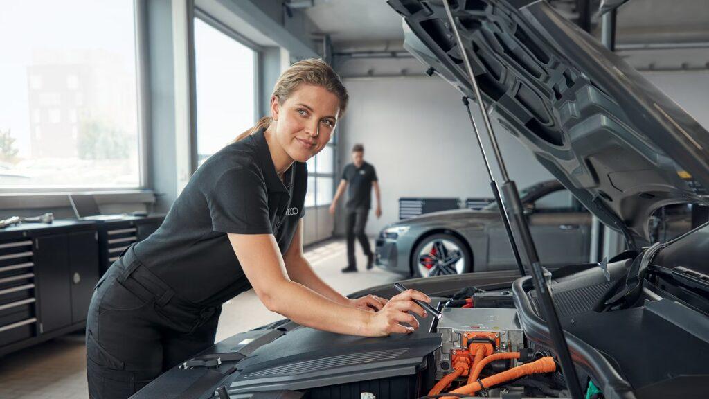 Mechanikerin steht vor einer geöffneten Motorhaube und leuchtet mit einer Taschenlampe in den Motorraum eines Autos