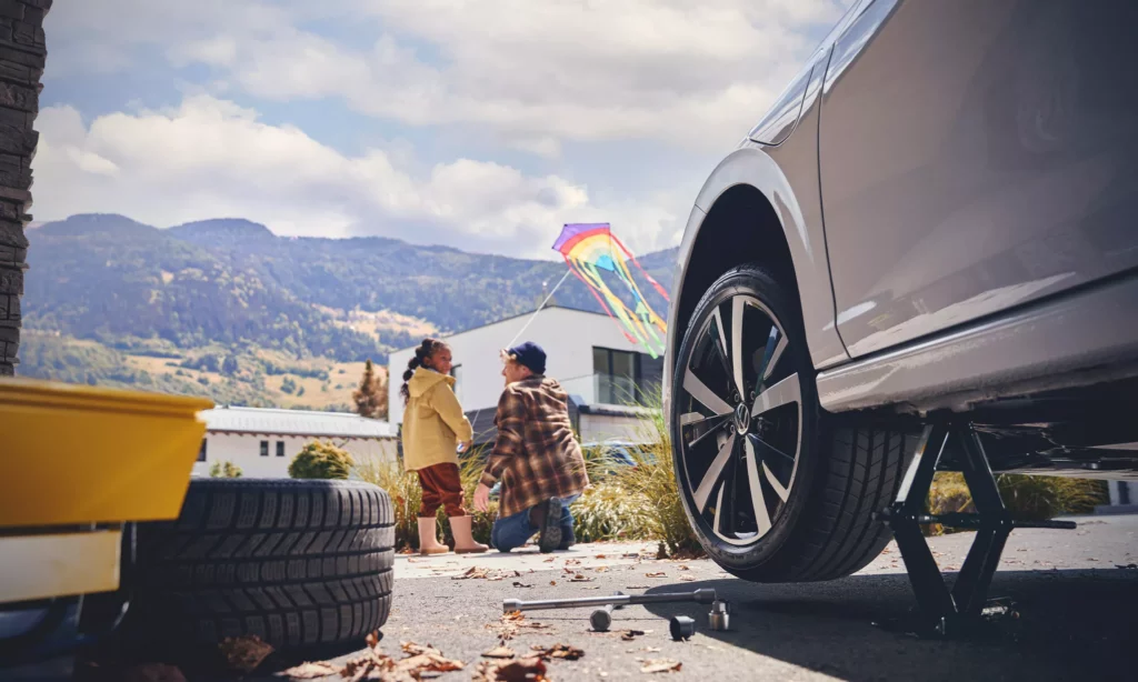 Vater kniet neben seiner Tochter, die einen gelben Regenmantel trägt und beide halten einen Drachen in der Hand, im Vordergrund ist der Reifen eines grauen VW Autos zu sehen