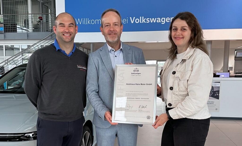 Drei Personen stehen in einem Autohaus vor einer blauen VW Willkommens-Wand, eine Person hält eine Urkunde, mit der Aufschrift 'Zertifikat Volkswagen Fleet Service'