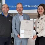 Drei Personen stehen in einem Autohaus vor einer blauen VW Willkommens-Wand, eine Person hält eine Urkunde, mit der Aufschrift 'Zertifikat Volkswagen Fleet Service'