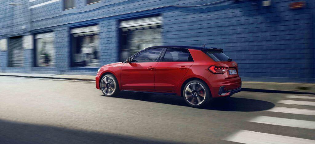 Roter Audi A1 Sportback fährt an einem blauen Gebäude vorbei, Seitenansicht, bei Tageslicht.