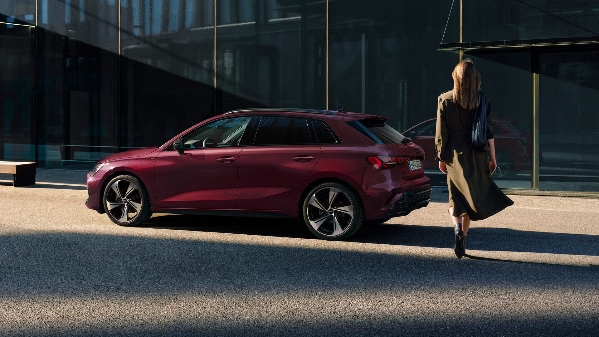 Roter Audi A3 Sportback steht vor modernem Glasgebäude, eine Frau mit langem Mantel und Tasche geht darauf zu
