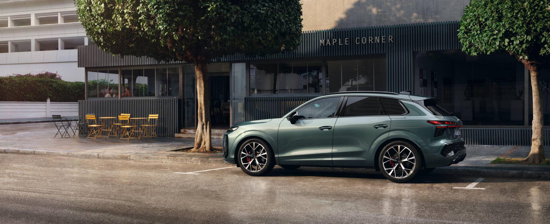 Seitenansicht des neuen Audi Q3 in grün-metallic, geparkt vor einem Café mit schwarzer Fassade und gelben Stühlen, umgeben von Bäumen.