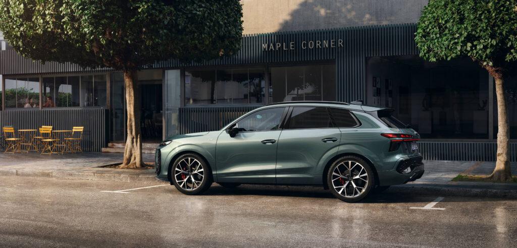 Markteinführung des neuen Audi Q3 2 Seitenansicht des neuen Audi Q3 in grün-metallic, geparkt vor einem Café mit schwarzer Fassade und gelben Stühlen, umgeben von Bäumen.