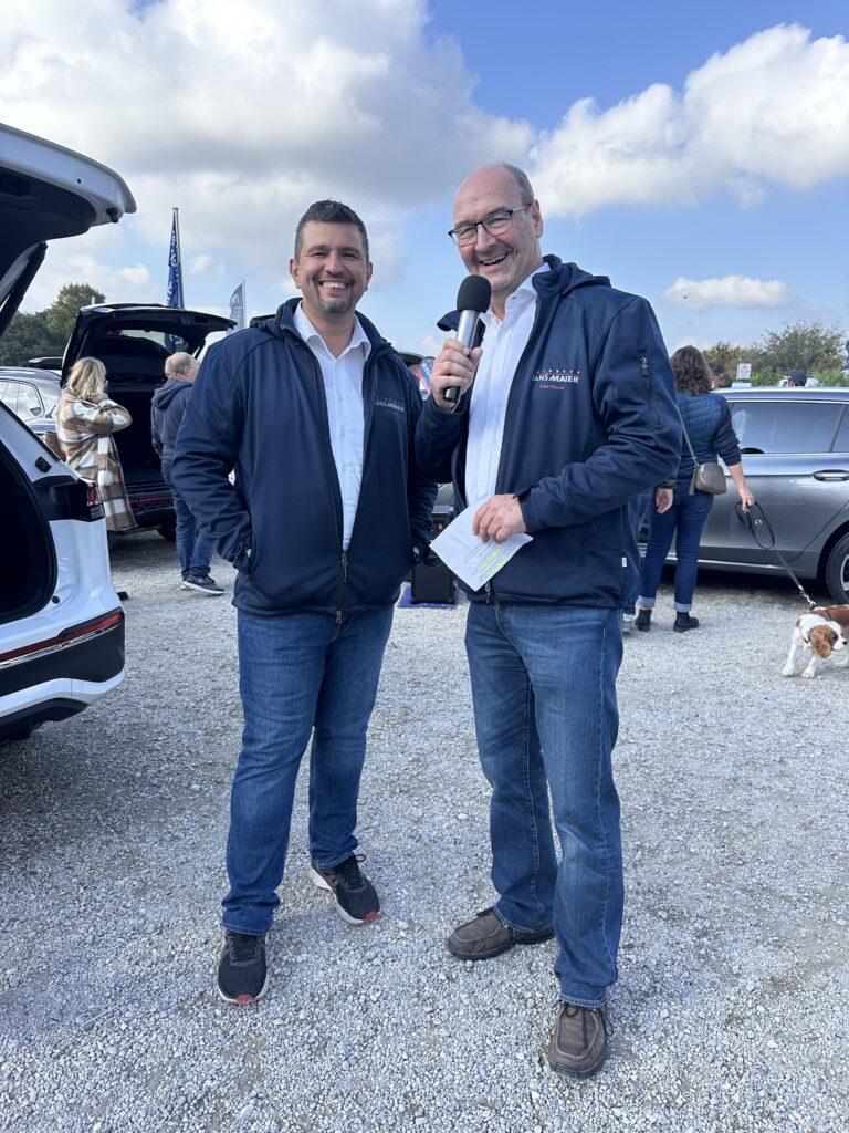 Großes Interesse an der Automobil Ausstellung Erding 2025 2 Zwei Männer in blauen Jacken mit weißem Hemd, einer hält Mikrofon und Notizzettel, stehen auf dem Volksfestplatz bei der Automobilausstellung Erding 2025.