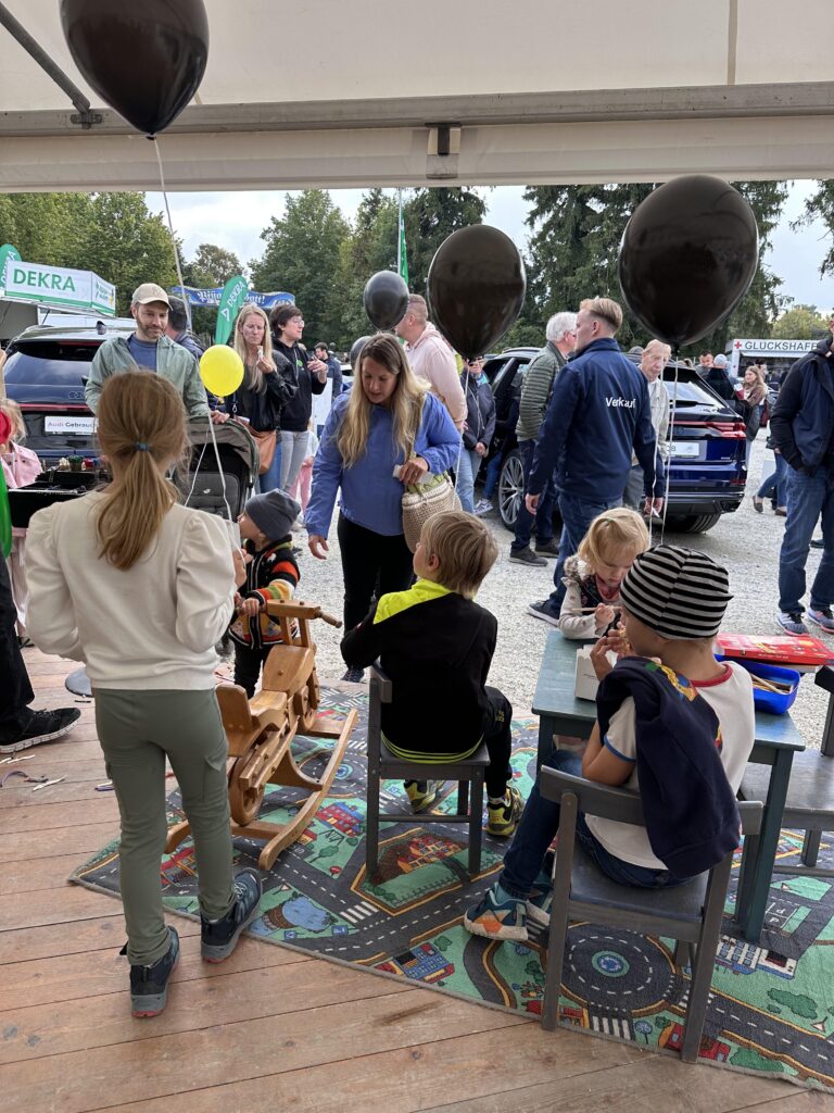 Großes Interesse an der Automobil Ausstellung Erding 2025 12 Kinder sitzen und stehen auf einem bunten Spielteppich mit Straßenmuster, umgeben von schwarzen und einem gelben Luftballons, im Hintergrund Menschen bei einer Automobilausstellung.