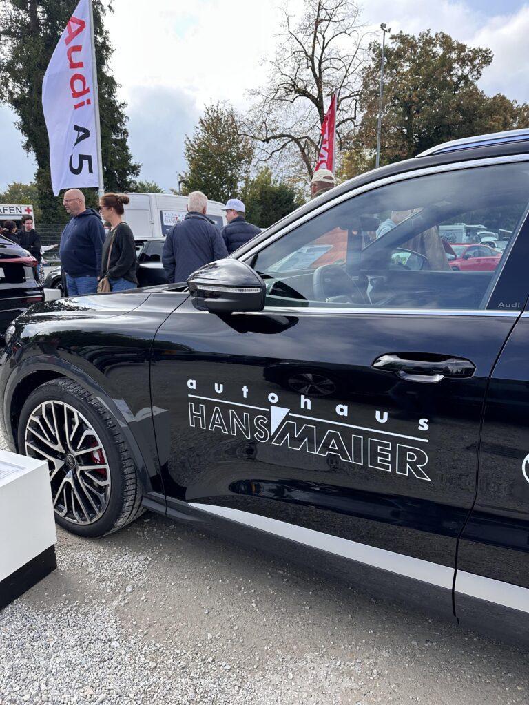 Großes Interesse an der Automobil Ausstellung Erding 2025 5 Schwarzer Audi SUV mit Aufschrift 'Autohaus Hans Maier' auf der Fahrertür, im Hintergrund Menschen und Audi-Fahnen auf der Automobilausstellung Erding 2025.