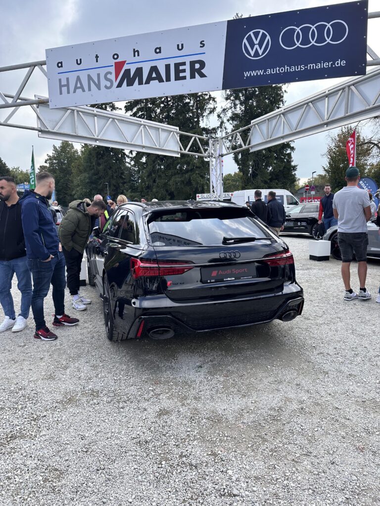 Großes Interesse an der Automobil Ausstellung Erding 2025 27 Schwarzes Audi Sport Auto mit vier Auspuffendrohren auf Kiesboden, umgeben von Menschen auf der Automobilausstellung Erding 2025, darunter Banner von Autohaus Hans Maier und Volkswagen sowie Audi-Logos.