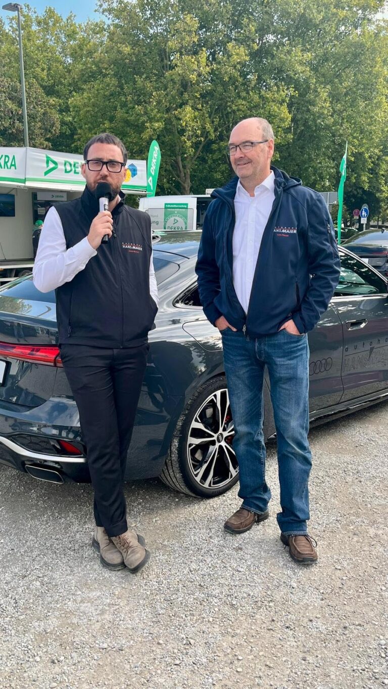 Großes Interesse an der Automobil Ausstellung Erding 2025 20 Zwei Männer in Jacken mit 'Autohaus Hans Maier' Logo bedruckt stehen vor einem dunkelgrauen Audi mit großen Felgen bei der Automobilausstellung Erding 2025, im Hintergrund Fahnen der Dekra zu sehen.