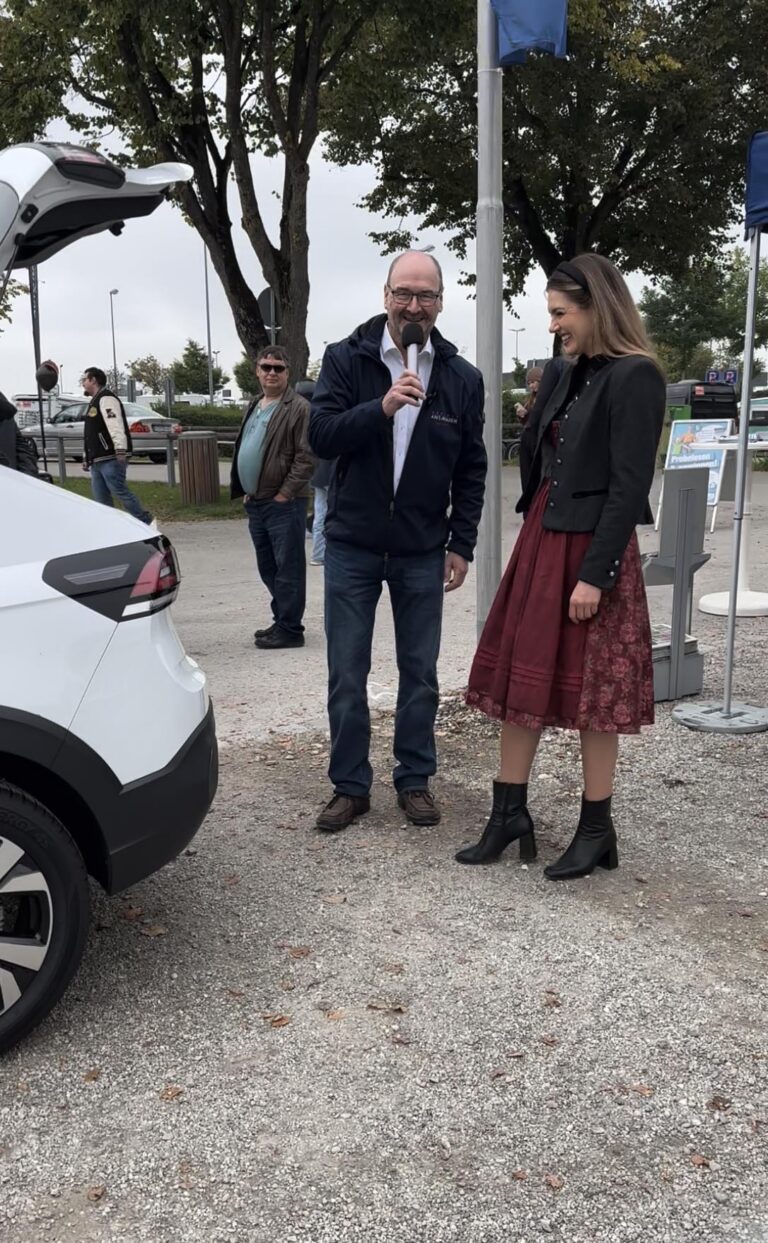 Großes Interesse an der Automobil Ausstellung Erding 2025 31 Mann mit Mikrofon und Frau in rotem Dirndl und schwarzer Trachten-Jacke stehen neben geöffnetem Kofferraum eines weißen Autos auf der Automobilausstellung Erding 2025.