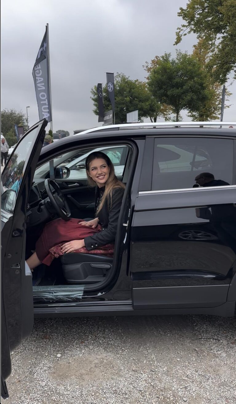 Großes Interesse an der Automobil Ausstellung Erding 2025 10 Herbstfestmadl 2025 sitzt auf Fahrersitz eines schwarzen Autos mit geöffneter Tür auf Automobilausstellung Erding 2025, im Hintergrund Fahnen und Bäume.