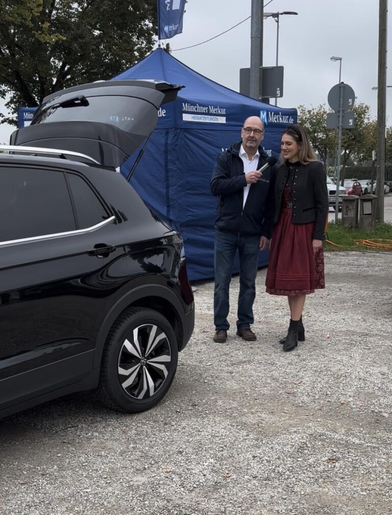 Großes Interesse an der Automobil Ausstellung Erding 2025 32 Schwarzes Auto mit geöffnetem Kofferraum auf Kiesplatz, zwei Personen stehen vor blauem Zelt mit Aufschrift 'Münchner Merkur' und 'Merkur.de'.