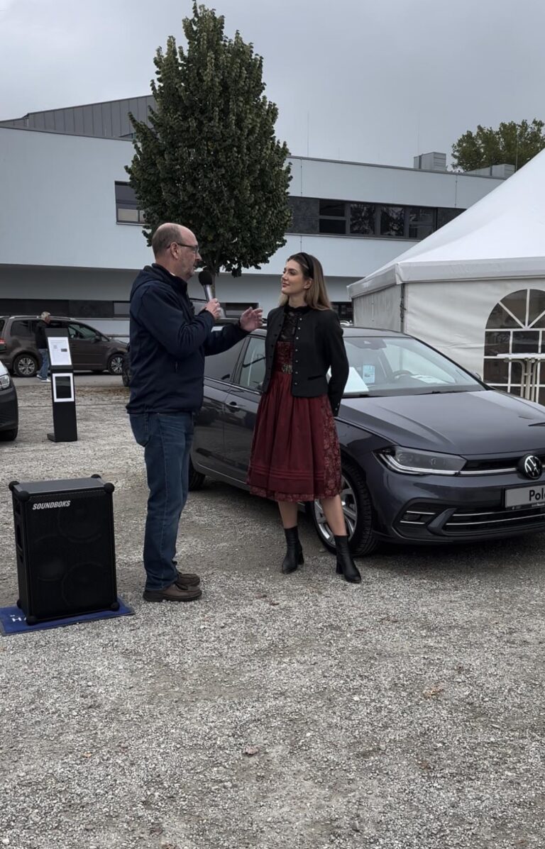 Großes Interesse an der Automobil Ausstellung Erding 2025 35 Mann mit Mikrofon spricht mit Frau in rotem Dirndl und schwarzer Jacke vor grauem Volkswagen Polo auf Automobilausstellung Erding 2025, Zelt und Baum im Hintergrund.