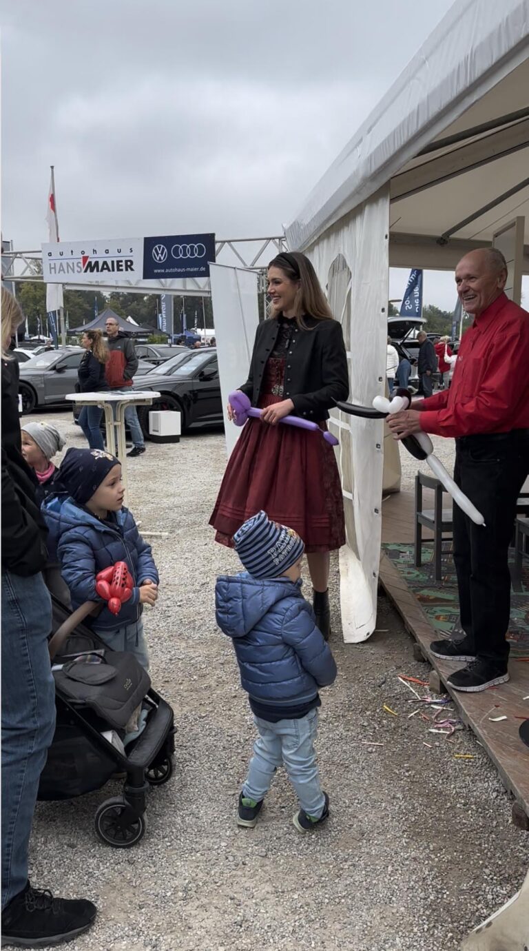 Großes Interesse an der Automobil Ausstellung Erding 2025 33 Frau in rotem Kleid und schwarzer Jacke hält lila Luftballonfigur, zwei Kinder in blauen Jacken stehen davor, Mann in rotem Hemd reicht weiße Luftballonfigur, Automobilausstellung im Hintergrund.