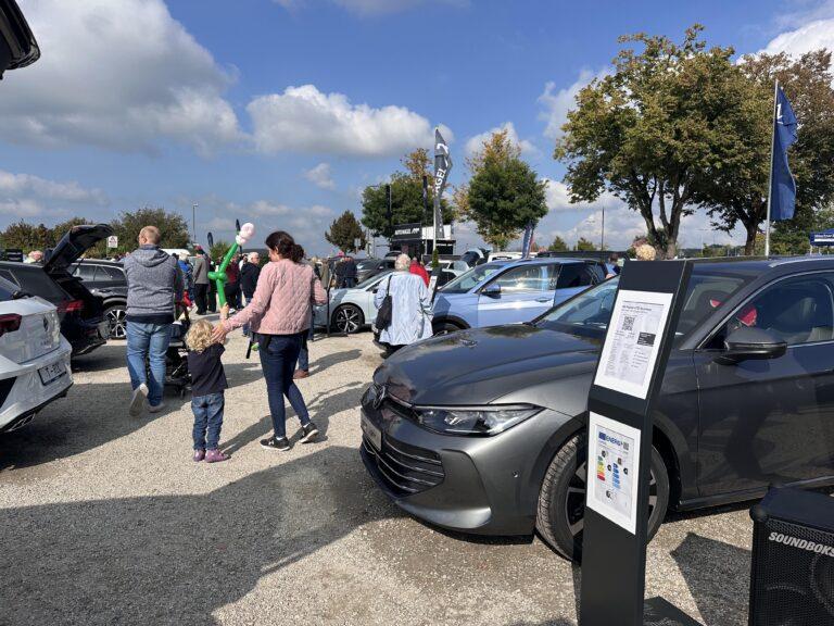 Großes Interesse an der Automobil Ausstellung Erding 2025 7 Mehrere Personen, darunter eine Frau mit rosa Jacke, gehen zwischen geparkten Autos auf einem Schotterplatz bei sonnigem Himmel. Ein graues VW Auto mit Informationsschild steht im Vordergrund.
