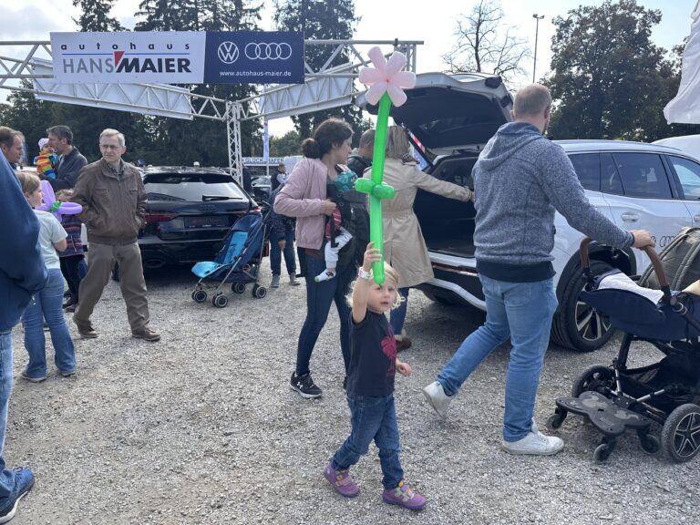 Großes Interesse an der Automobil Ausstellung Erding 2025 8 Kind hält grünen Luftballon in Form einer Blume, umgeben von Besuchern auf dem Volksfestplatz Erding bei der Automobilausstellung vor Autohaus Hans Maier mit VW- und Audi-Logo.
