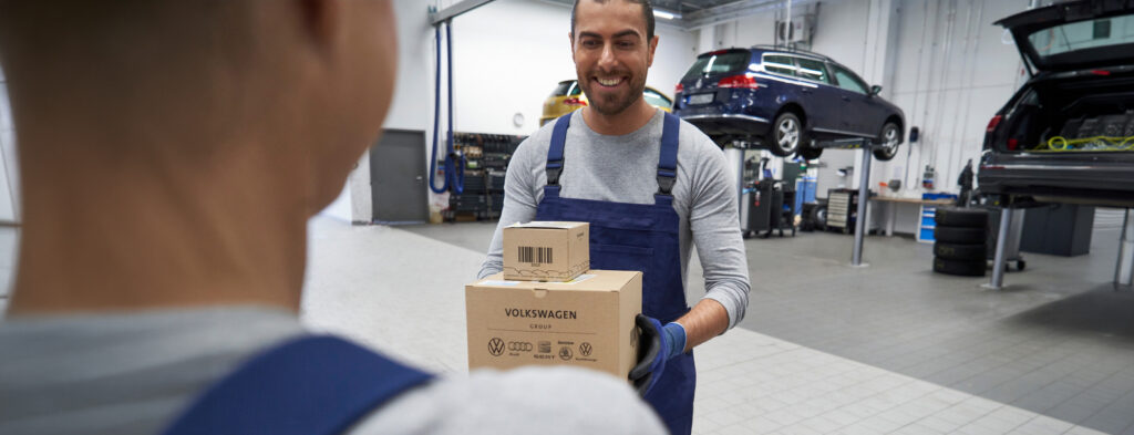 Zwei Männer in blauer Arbeitskleidung in einer Autowerkstatt, einer hält zwei Kartons mit Volkswagen-Logos