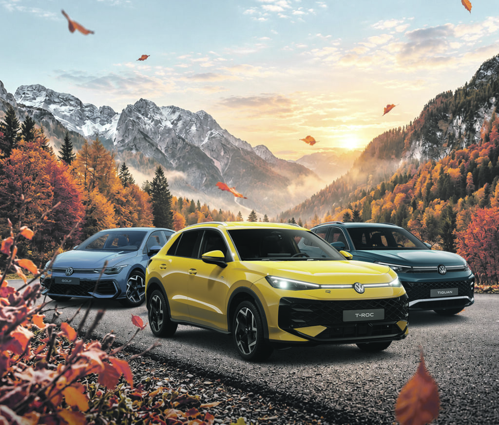 Drei Autos fahren auf einer Bergstraße mit herbstlich gefärbtem Wald und Bergen im Hintergrund