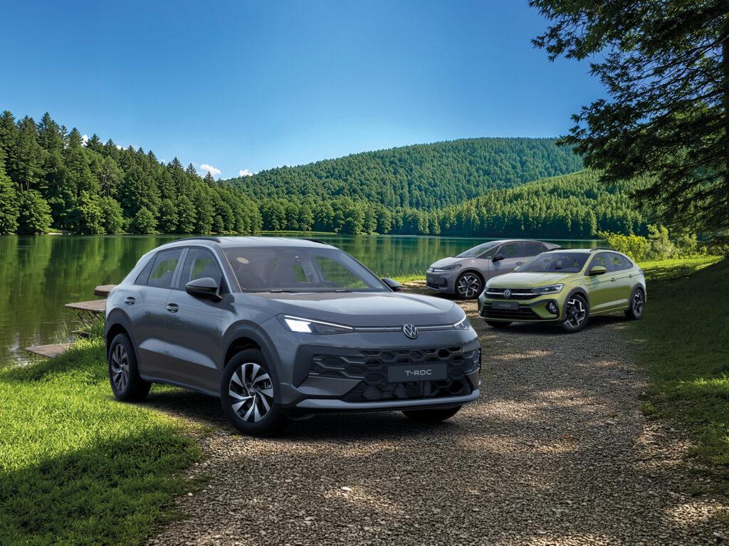 Drei Volkswagen-SUVs, vorne ein grauer T-Roc, dahinter ein Tiguan und ein weiteres Modell, geparkt an einem See mit bewaldeten Hügeln im Hintergrund