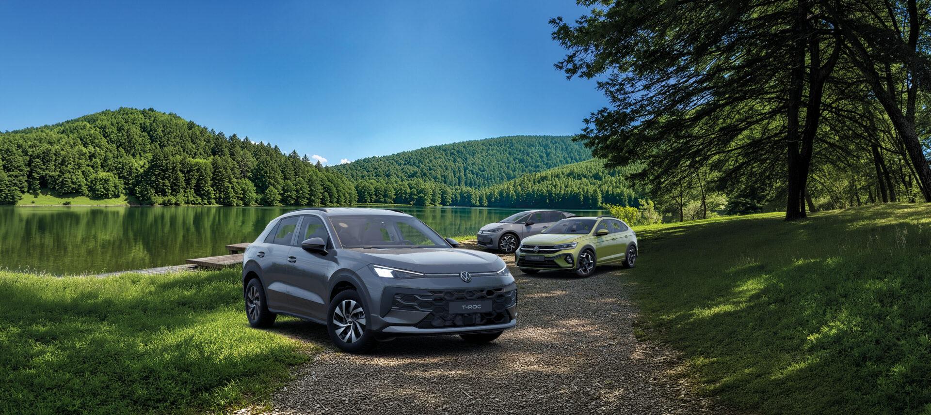 Dein VW Sommer startet jetzt! 1 Drei Volkswagen-SUVs, vorne ein grauer T-Roc, dahinter ein Tiguan und ein weiteres Modell, geparkt an einem See mit bewaldeten Hügeln im Hintergrund