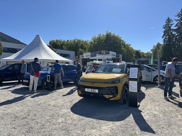 Gelber Volkswagen T-Roc steht auf einem Schotterplatz bei einer Autoschau mit mehreren Besuchern und weißen Zelten im Hintergrund