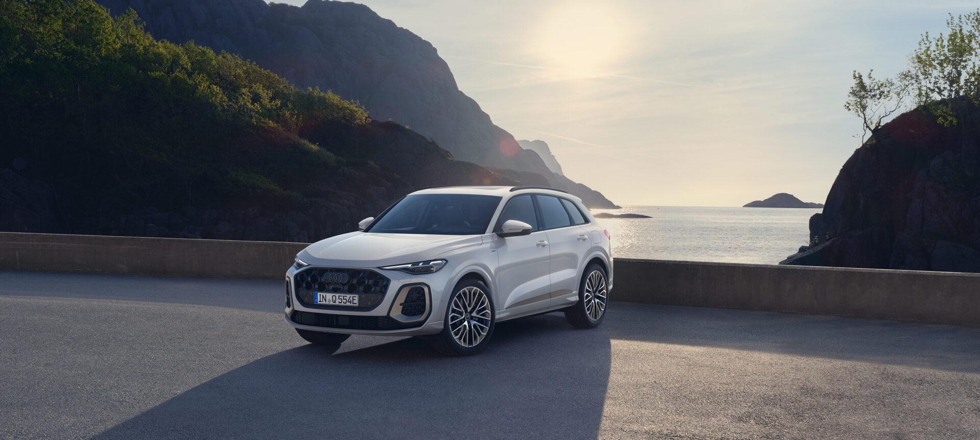 Weißer Audi SUV steht auf einer Küstenstraße mit Berglandschaft im Hintergrund bei Sonnenuntergang